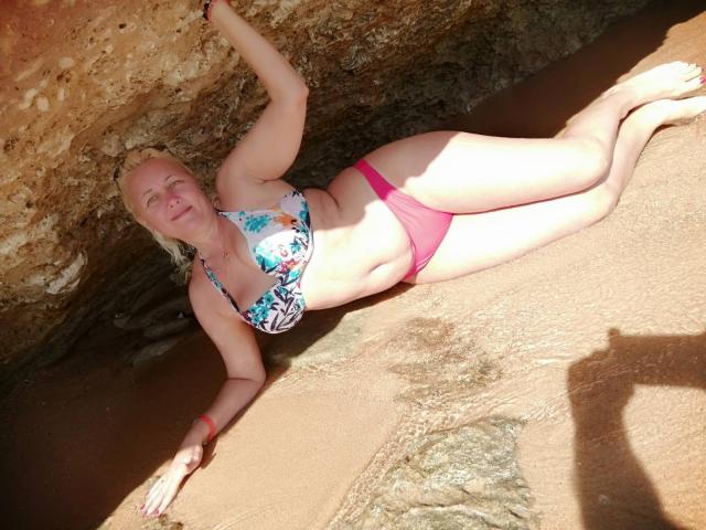 CaronCapella, eine blonde Frau, liegt auf einem sandigen Strand, trägt einen bunten Bikini-Oberteil und einen pinken Bikini-Unterteil, der rechte Arm ist angehoben, ein Schatten einer Person ist rechts zu sehen, sonnendurchflutet, felsiger Hintergrund einer Höhle..