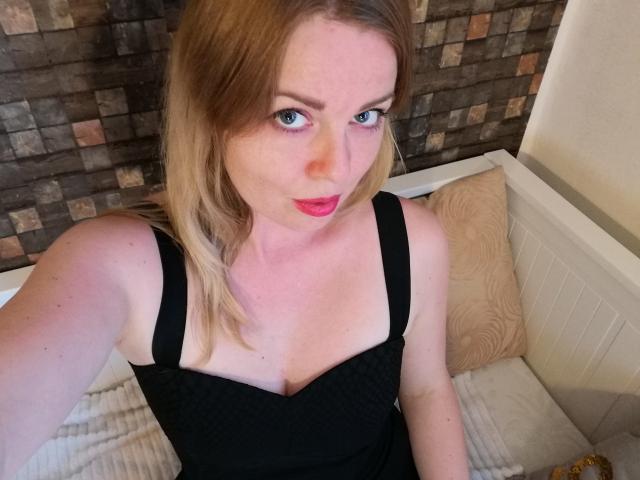 Nicole Linharts, con cabello rubio, ojos azules y lápiz labial rojo, vestida con un vestido negro que deja ver sus senos, sentada en una cama con un cojín beige..