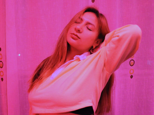 AradiaX with pink-tinted skin, long red hair, eyes closed, wearing a white shirt, arms raised, showing her tits, gold hoop earrings, soft pink glow, relaxed expression.