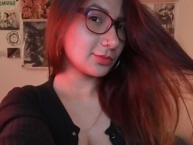 AradiaX with glasses, nose ring, and hoop earrings, has long brown hair, red lighting, black shirt, and a slight smile, showing her tits. She's looking at the camera.