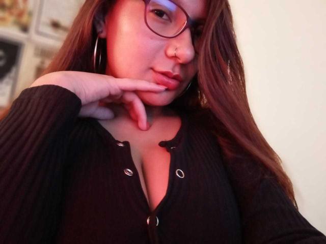 AradiaX con gafas, aro en la nariz y cabello largo castaño, vestida con una camiseta negra acanalada, con la mano en la barbilla, mostrando sus pechos, una luz rosa suave en su rostro..