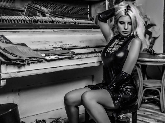 GoddessKaterina, com cabelo loiro, vestindo um vestido preto de látex brilhante, com amarração frontal e luvas longas, sentada em uma cadeira, com as mãos no cabelo, aparência sexy, seios grandes, fundo com um piano..