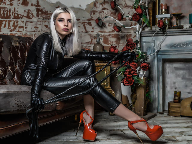 GoddessKaterina with long white hair, black leather outfit, and red high heels, holding a black whip, sitting on a brown couch, surrounded by red roses.