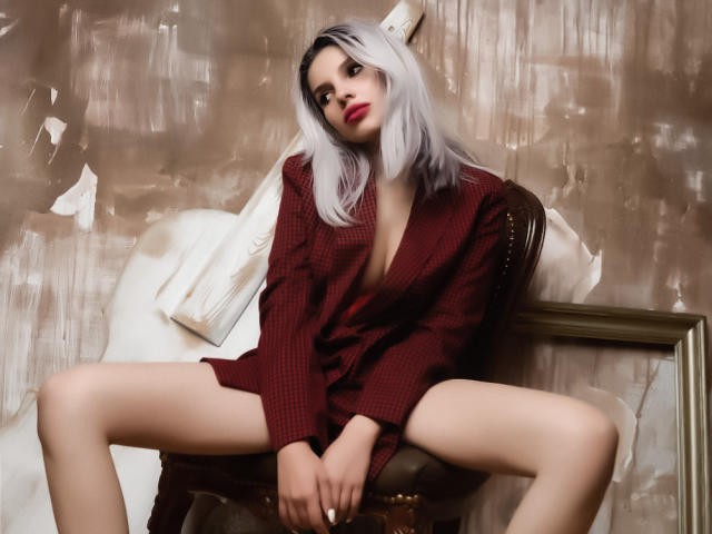 GoddessKaterina, con piel pálida, cabello plateado, labios rojos y una camisa de cuadros rojo oscuro, con las piernas separadas, sentada en una silla, mostrando su vulva, con las manos sobre sus muslos..