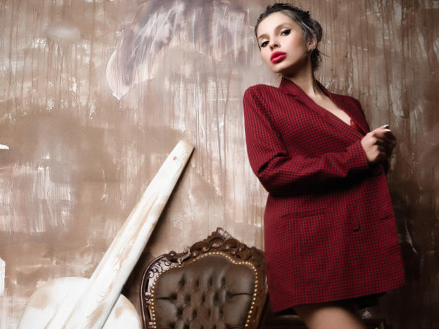 GoddessKaterina with red checkered blazer, standing, hands near chest, confident, sexy, dark hair, red lipstick, wooden chair with tufted leather, and white wooden panel in background.