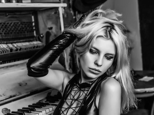 GoddessKaterina with blonde hair, wearing a black lace-up top and shiny black gloves, looking down with a slightly pouty expression, standing by an old, dusty piano.