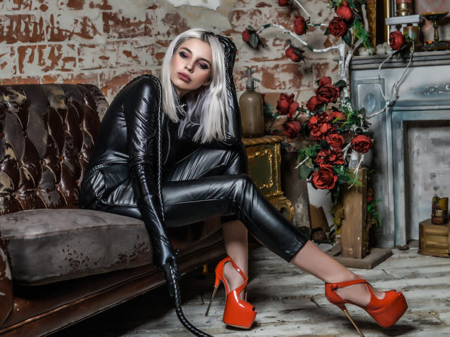 GoddessKaterina, con i capelli argentati, un abito in lattice nero e tacchi alti rossi, seduta su un divano marrone sporco, che tiene in mano una frusta nera, con un aspetto sexy, con rose rosse sullo sfondo..