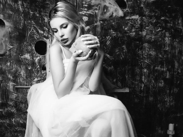GoddessKaterina with blonde hair, wearing a white dress, sits holding a glass, her hands on her small tits, in a black-and-white, textured, gritty photo.
