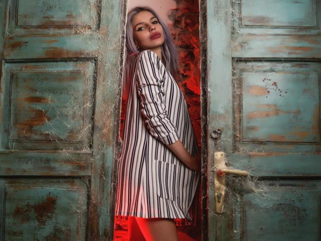 GoddessKaterina with long purple hair, striped black and white dress, red skirt, standing in a worn, green, rustic wooden door, looking over shoulder with a sultry expression.