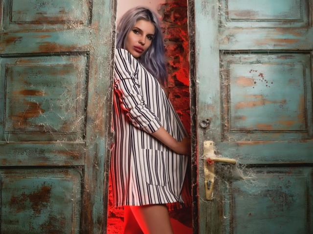 GoddessKaterina, avec ses cheveux violets, sa robe rayée et sa culotte rouge, se tient devant une porte rustique, bleu-vert et patinée, et regarde séductrice l'appareil photo avec un léger sourire..