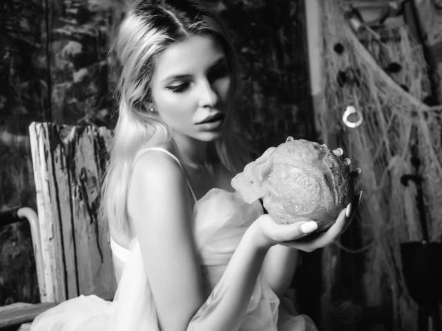 GoddessKaterina with long blonde hair, wearing a white dress, holding a textured rock in her hands, looking down with a slightly pouty expression, in a black-and-white photo.