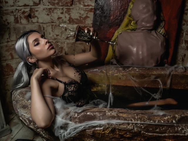 GoddessKaterina, con cabello plateado, blusa de encaje, vertiendo agua sobre sus senos desde un grifo de metal, sentada en una bañera oxidada llena de agua, rodeada de vapor y gotas de agua..