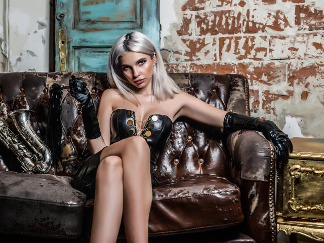 GoddessKaterina with silver hair, black leather corset, and gloves, sitting on a brown, tufted leather couch, legs crossed, showing her tits and a seductive, serious expression.