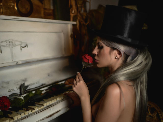GoddessKaterina with long silver hair, topless, wearing a black top hat, holding a red rose to her lips, sitting at an old, white piano with roses on the keys.