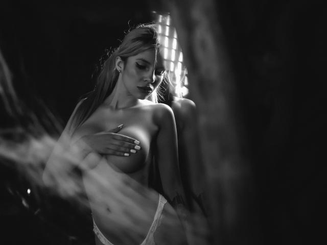 GoddessKaterina with long blonde hair, covering her tits with one hand, wearing lace panties, standing in dark, moody black-and-white shot, looking down with soft, closed eyes.