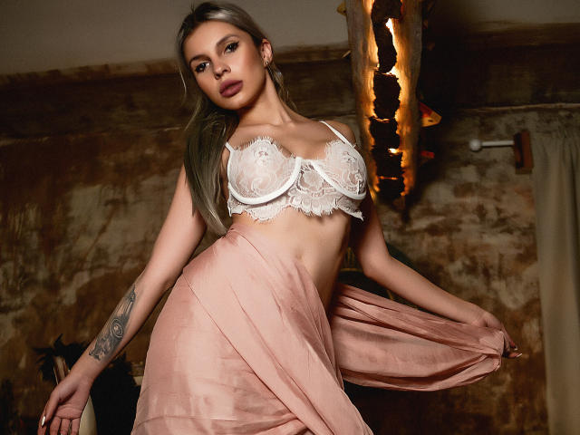 GoddessKaterina with long blonde hair, wearing a white lace bralette showing her tits and a pink sheer skirt, tattoo on right arm, standing in a dimly lit room.