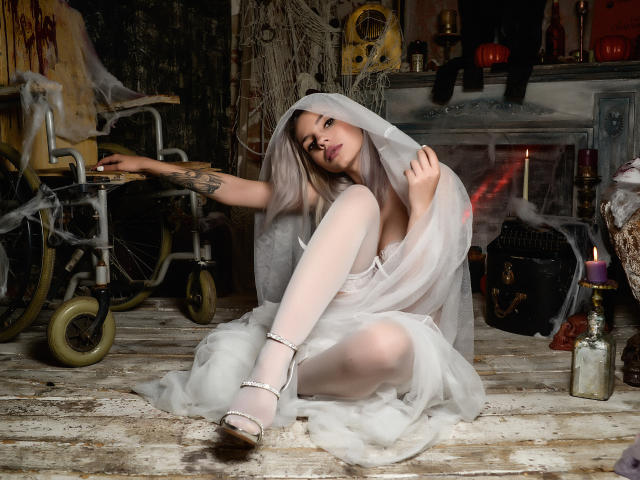 GoddessKaterina com meias brancas, véu transparente e saltos altos, sentada em um chão de madeira, mostrando seus seios, com uma tatuagem no braço esquerdo e um olhar sensual..