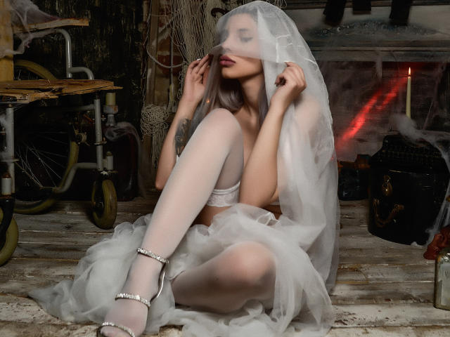 GoddessKaterina with pale skin, white lingerie, and a sheer veil, sitting on a wooden floor, legs crossed, white stockings, and high heels, holding the veil near her face.