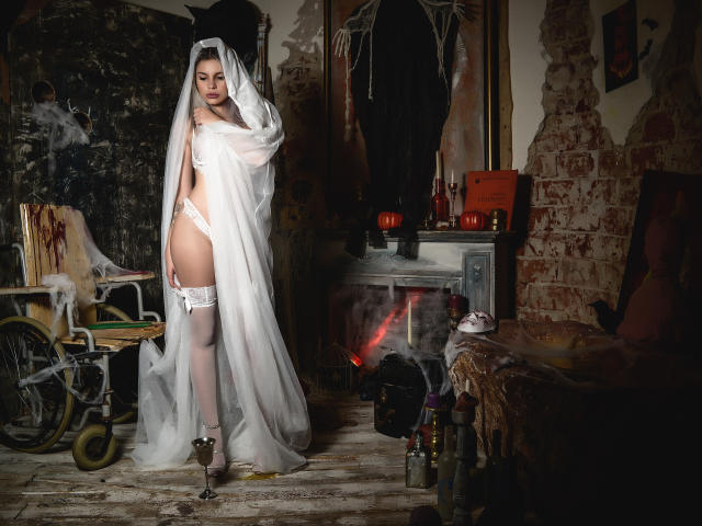 GoddessKaterina with white veil, lace lingerie, garter, and stockings, standing in dim room, wearing high heels, showing her ass, and looking seductive with a slight smile.