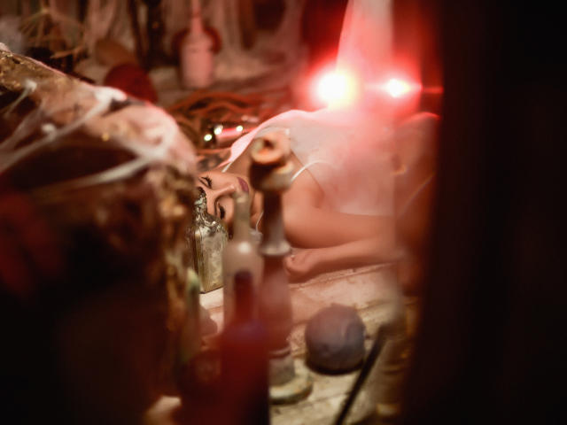 GoddessKaterina's bare ass is visible as she lies on a rustic, cluttered surface with a red light flare in the background, surrounded by various bottles and objects.