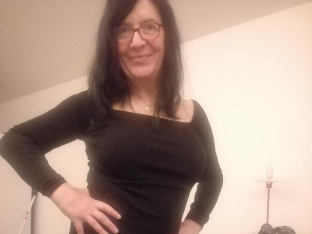 Emilianne with black hair, glasses, and a black long-sleeve top, showing off her tits and smiling with hands on hips. She's wearing a gold necklace.