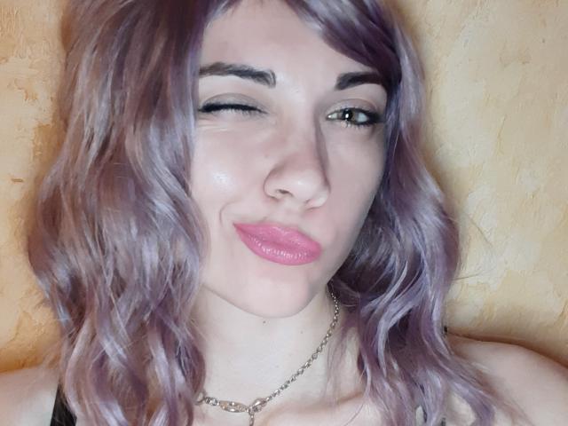 BethAh a les cheveux ondulés d'un violet clair, elle cligne un œil et a les lèvres roses et brillantes. Elle porte un haut noir et un collier en argent..
