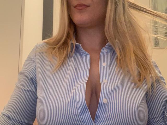 QueenKaly with blonde hair, wearing a blue-striped button-up shirt, slightly open to show her cleavage and small tits. Her lips are gently closed.