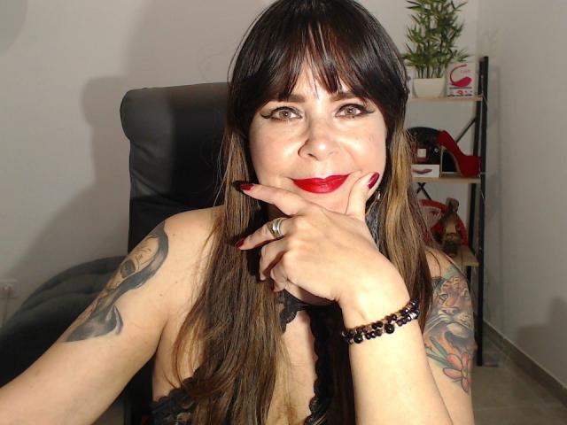EvaLuv with long brown hair, red lipstick, tattoos on her arms, black lace top, red nails, ring on finger, black bracelet, hand on chin, smiling, black chair behind.