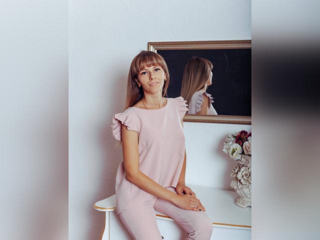 Sophie Wiles, con cabello castaño largo, una blusa rosa con volantes y pantalones rosas, sentada en una silla blanca, reflejo en el espejo, un ramo de flores a la derecha..