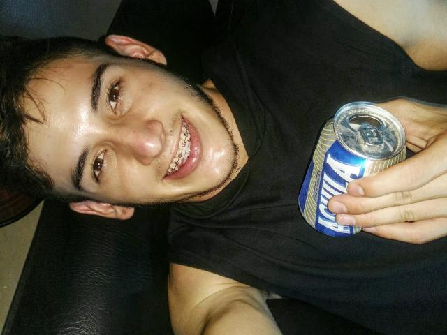 JonathanJeremiah with a black shirt, holding a can of beer, smiling, showing slightly gapped teeth, and a short beard, lying on a dark leather surface.