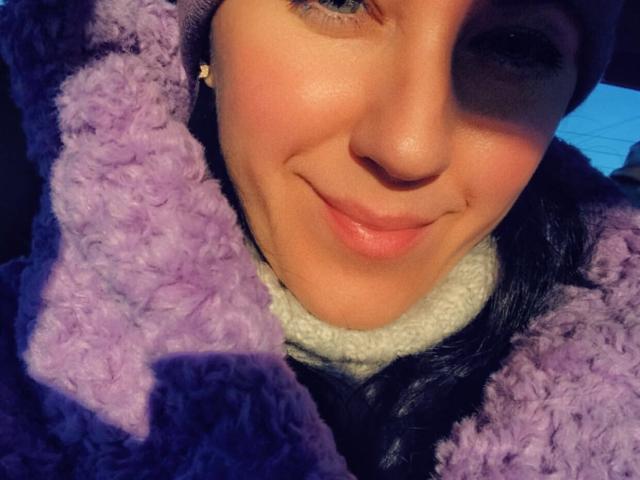 Misstyk's face, smiling slightly with purple, fuzzy, textured coat collar, and fair skin, wearing a brown beanie, blue eyes, and small gold earrings, in bright sunlight.