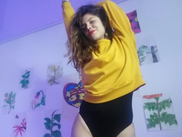 CeibaX, con cabello largo y castaño, vestida con una sudadera amarilla y tanga negra de corte alto, con los brazos levantados, sonriendo y mostrando su trasero. Está de pie, luciendo segura de sí misma, en una habitación colorida..