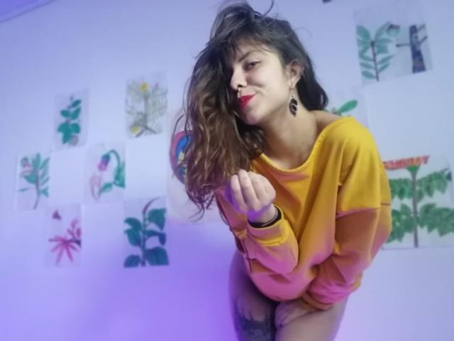 CeibaX, com cabelo castanho comprido, batom vermelho, suéter amarelo e brincos, curvada para frente, mostrando o bumbum, em um quarto com desenhos botânicos nas paredes..