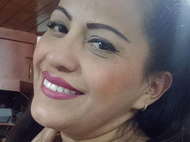 MelissaDelici's face, smiling with pink lipstick, dark eyeliner, and brown eyebrows. She's wearing a gold earring, and her dark hair is pulled back. Her nose is slightly upturned.