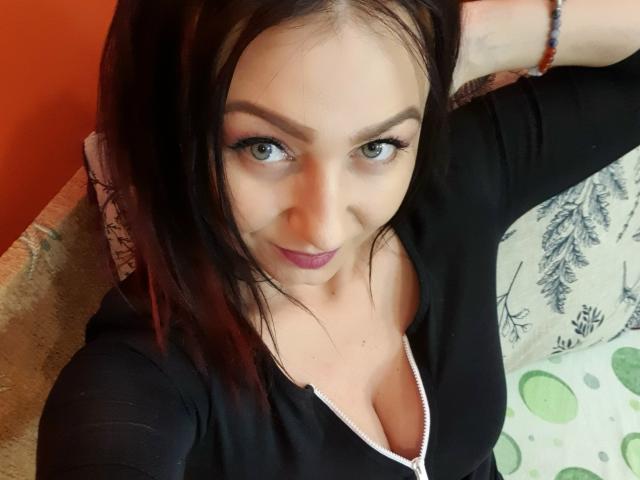 JolieXKitty with medium brown hair, green eyes, and pink lipstick, wearing a black top with a white zipper, showing her tits, smiling at the camera, arm raised.