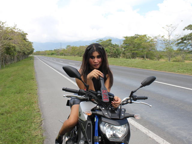 Samanta, con lunghi capelli castani e pelle abbronzata, sta guidando una motocicletta nera su una strada, tenendo il manubrio con la mano destra, indossando un vestito bianco e stivali neri..