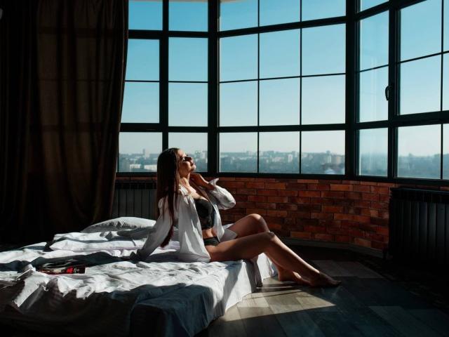 ViollinaLove, with long brown hair, sits on a bed in a sunlit room, wearing an open white shirt and black underwear, her tits visible, looking at a phone.