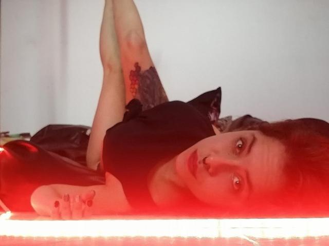 CeibaX está acostada sobre una superficie iluminada con luz roja, con las piernas levantadas, vistiendo una camiseta negra, con un piercing en la nariz, tatuajes en los brazos y cabello corto castaño, mirando a la cámara..