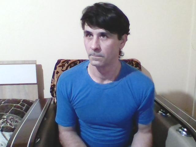 BlueJackie, con cabello negro corto, vistiendo una camisa azul ajustada, sentada en una cama con un cabecero estampado, mirando ligeramente hacia un lado, mostrando una expresión neutra..