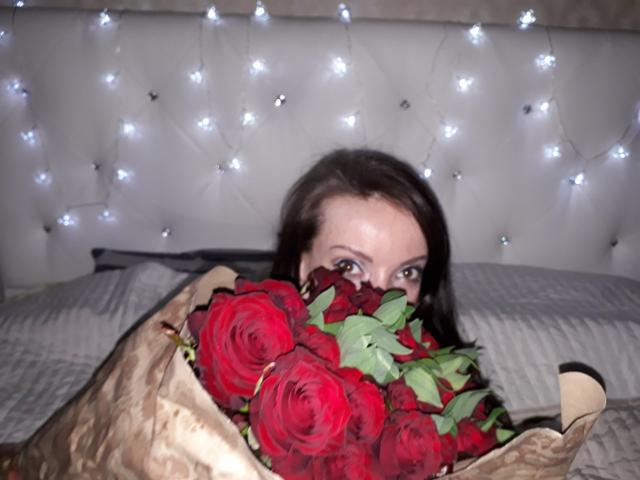 DonnaLaViva mit roten Rosen, die in braunem Papier ihre Brüste bedecken, ihre Augen schauen hervor, weiße Lichterketten im Hintergrund, auf einem grauen Bett..