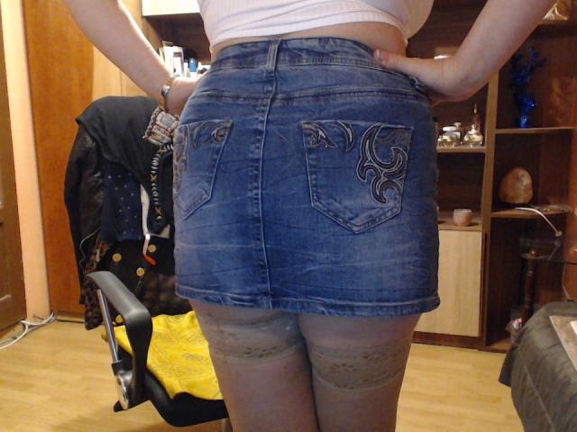 La gonna di jeans blu di LadyEvelynn, che mette in mostra il suo sedere, con tasche decorative, e calze grigie in pizzo, in piedi con le mani sui fianchi, in una stanza con mobili in legno..