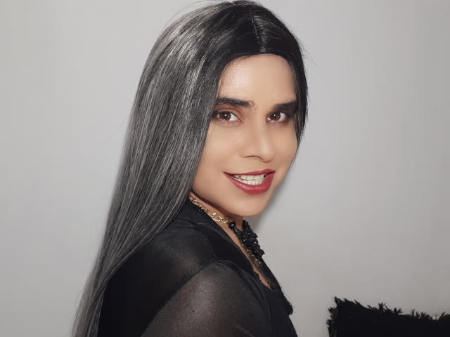 CamilaFetishDirty has long, sleek black hair, brown eyes, and red lipstick. She's smiling with visible teeth, wearing a black sheer top and a gold necklace.