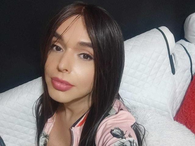 Jhovanna, con i capelli castano scuro, le labbra rosa e la pelle chiara, indossa una giacca fiorata rosa e guarda la fotocamera con un leggero sorriso, sdraiata su un cuscino bianco trapuntato..