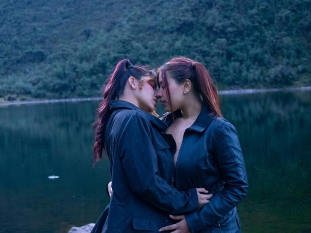 KorinYAmbar with red ponytails, dark jackets, and earrings, nose to nose, hands on each other's backs, standing by a calm lake, touching foreheads, intimate, dusk.