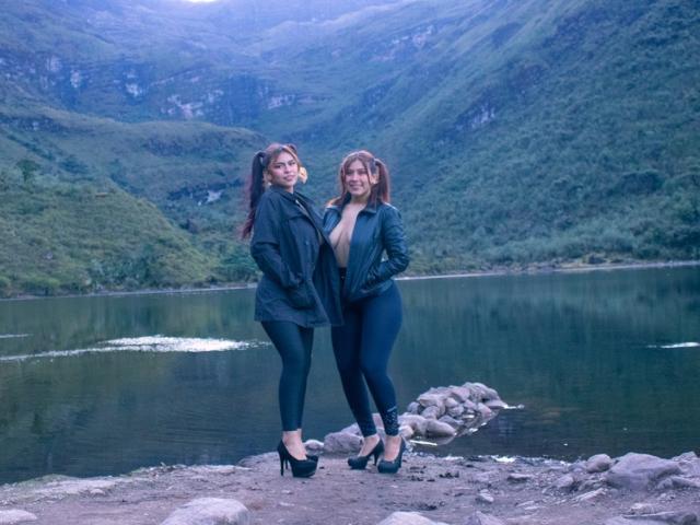KorinYAmbar stands with another KorinYAmbar by a lake, both in black outfits and high heels, with green mountains and a calm water backdrop, smiling at the camera.