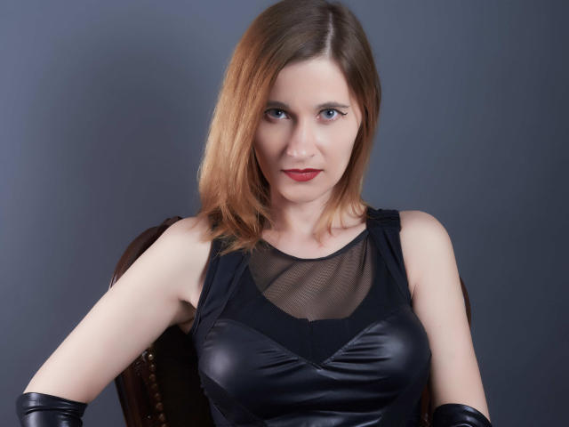 MistressKate, con un seno di media grandezza, indossa una maglia nera trasparente e guanti di pelle, ha i capelli biondi a metà schiena, rossetto rosso e un'espressione seria, ed è seduta contro uno sfondo scuro..