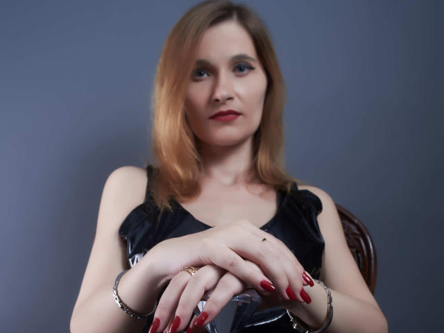 MistressKate with shoulder-length blonde hair, red lipstick, black dress, red nails, and silver bracelets, sitting with hands crossed, looking forward. Boobs slightly visible, but not the focus.