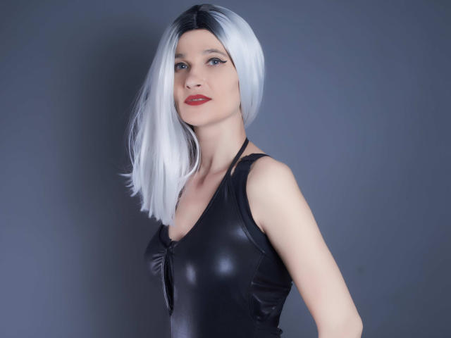 MistressKate with white bob, black leather top, red lipstick, pale skin, and a slight smirk, standing against a gray background. Boobs are covered by the top.