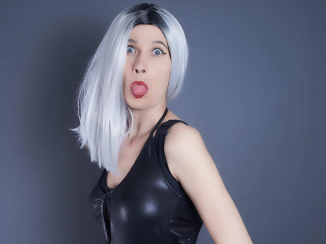 MistressKate with silver hair, pouty red lips, blue eyes, wearing black leather top, looking at camera, slightly turned, grey background, medium shot, soft light, slight shadow.