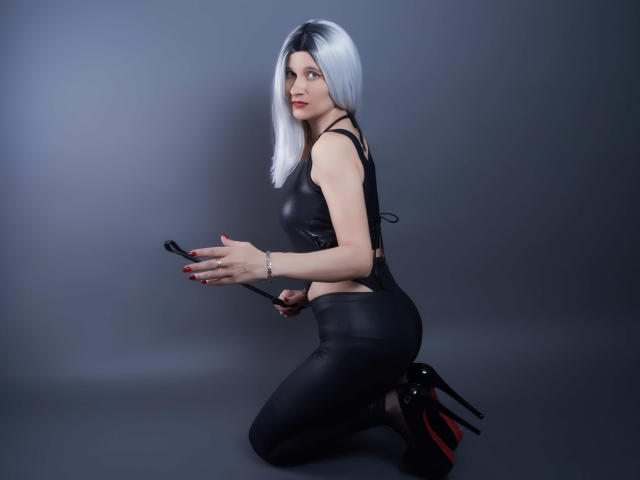 MistressKate with silver bob, black leather top, and pants, holding a whip, kneeling, wearing black high heels with red soles, showing off her ass, with red nails.